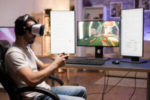A gamer wearing a VR headset sits at a desk with three screens, actively engaged in gaming and managing chat.