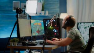 A gamer sits at a desk with dual monitors, a microphone, and gaming equipment, focused on gameplay in a well-lit room.