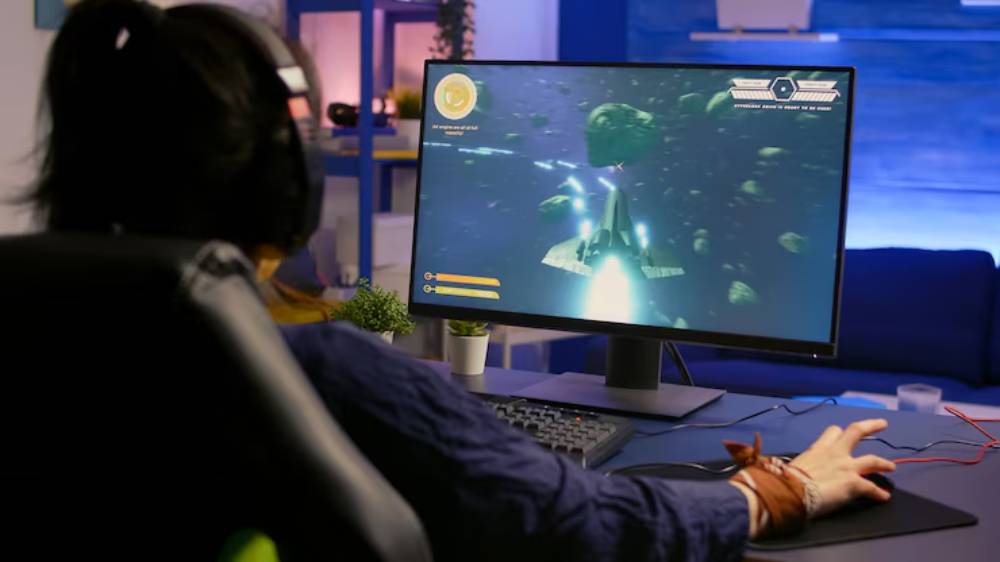 A gamer wearing headphones focuses on a computer screen displaying a space-themed video game, navigating through asteroids.