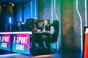 A gamer seated at a desk with gaming equipment, focused on the computer in a brightly lit e-sports arena.