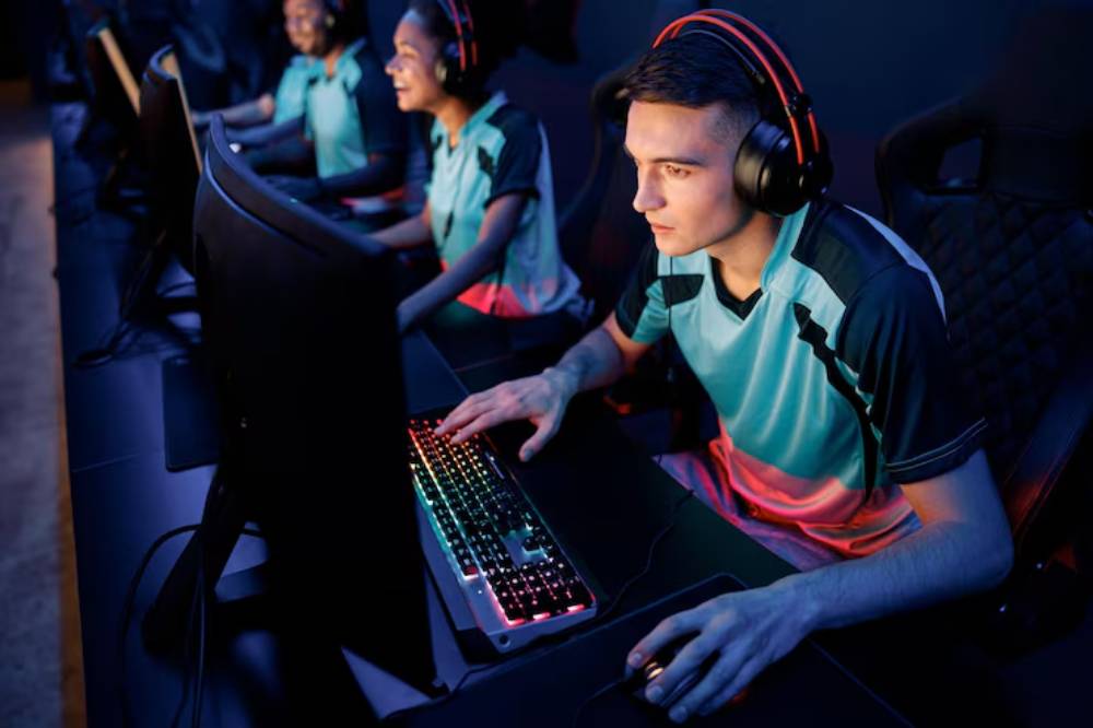 Gamers in matching teal jerseys use gaming computers in a dark, modern esports setup with colorful LED lighting.