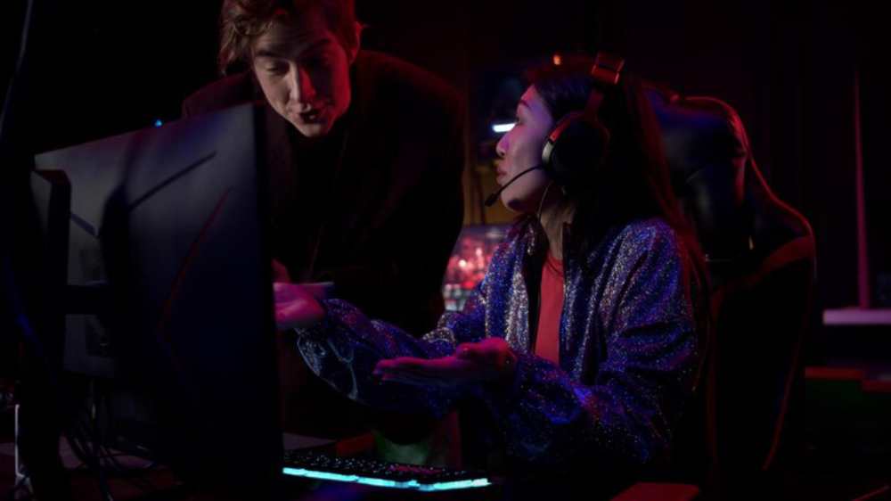 A gamer wearing headphones gestures animatedly while seated at a computer, surrounded by colorful lighting in a gaming setup.