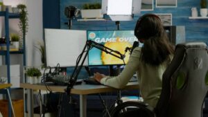 A gamer in headphones sits at a desk with dual monitors, featuring a "Game Over" screen, surrounded by studio equipment and plants.