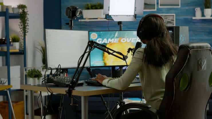 A gamer in headphones sits at a desk with dual monitors, featuring a "Game Over" screen, surrounded by studio equipment and plants.