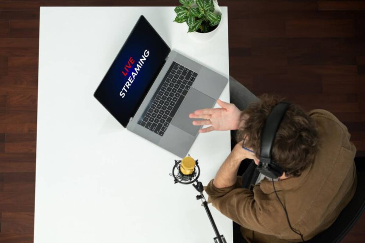 A person in headphones gestures while live streaming on a laptop, with a microphone and a potted plant on a white desk.