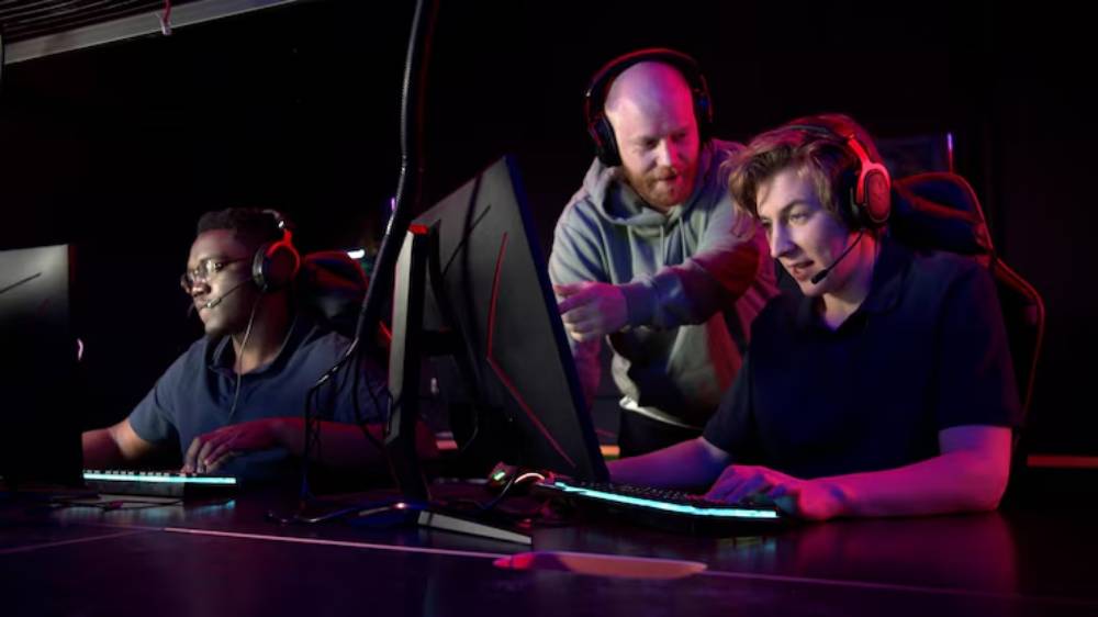 A group of gamers sit in a darkened room and their coach is mentoring them, they are wearing headsets and engaged in gameplay.