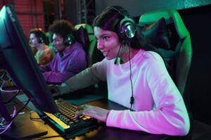 A gamer wearing headphones focuses intently on the keyboard and monitor in a dimly lit esports environment with vibrant lights.