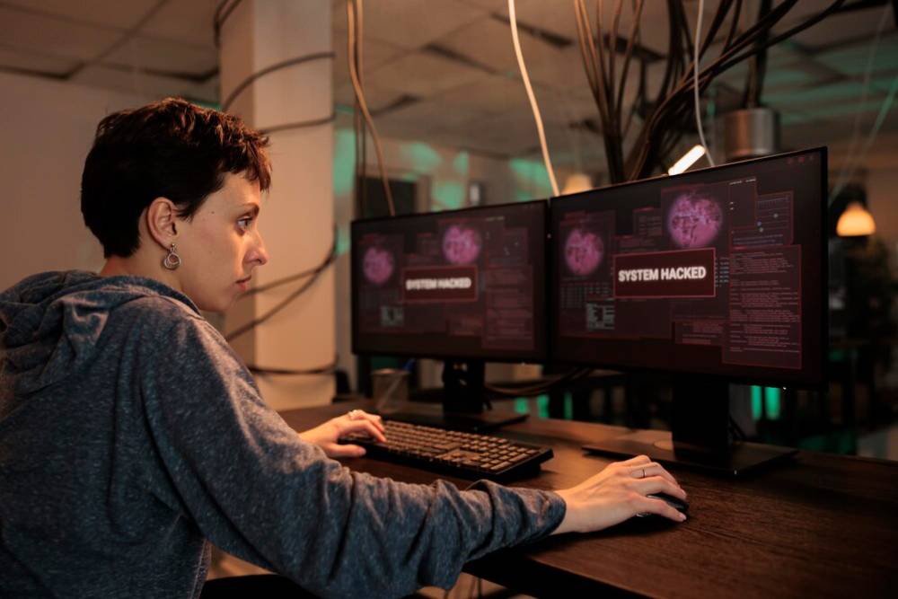 A person sits at a desk with two monitors displaying hacking alerts, including 