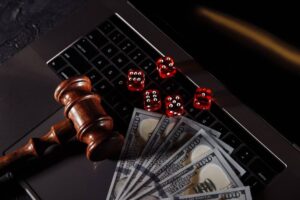 A wooden gavel rests on a laptop keyboard, surrounded by red dice and fanned-out hundred-dollar bills, suggesting gambling themes.