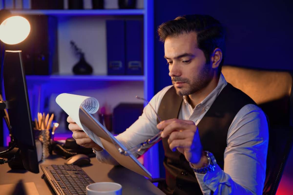 A person in formal attire sits at a desk, holding a rolled document and glasses, with a warm desk lamp and blue-lit shelves in the background.