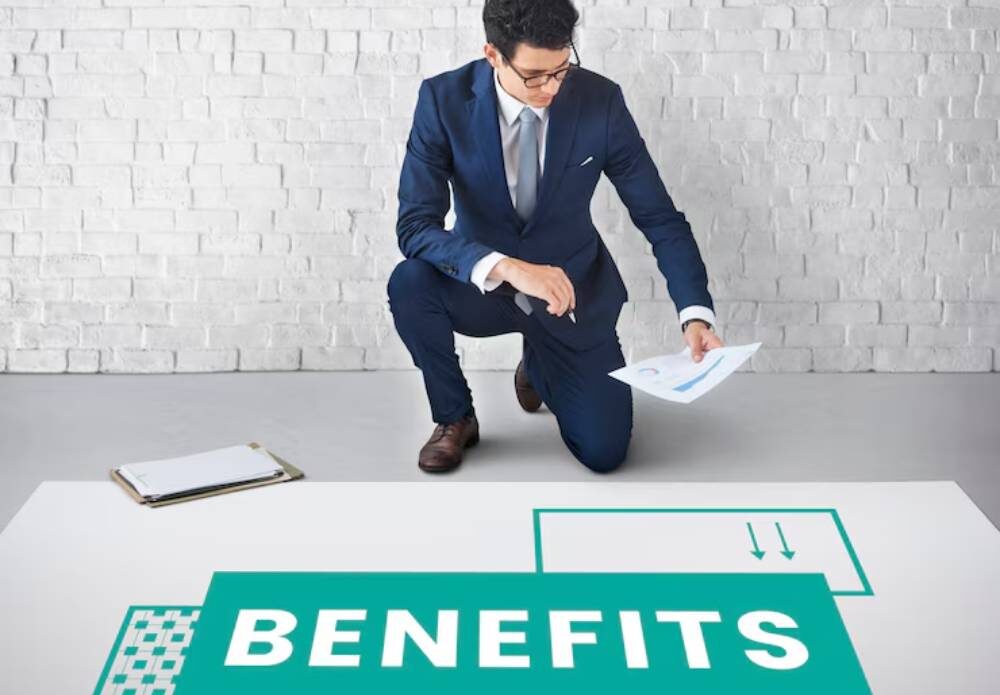 A businessman in a blue suit kneels beside a floor graphic that prominently displays the word 'BENEFITS'.