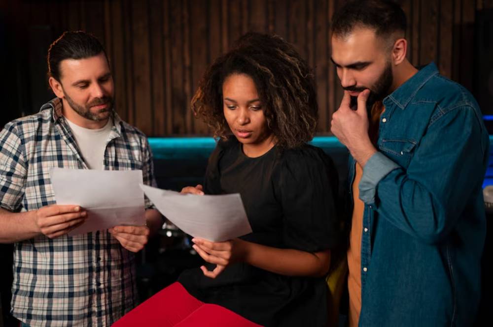 Three individuals gather around a table, focused on sheets of paper, engaged in discussion in a dimly lit, music-themed environment.
