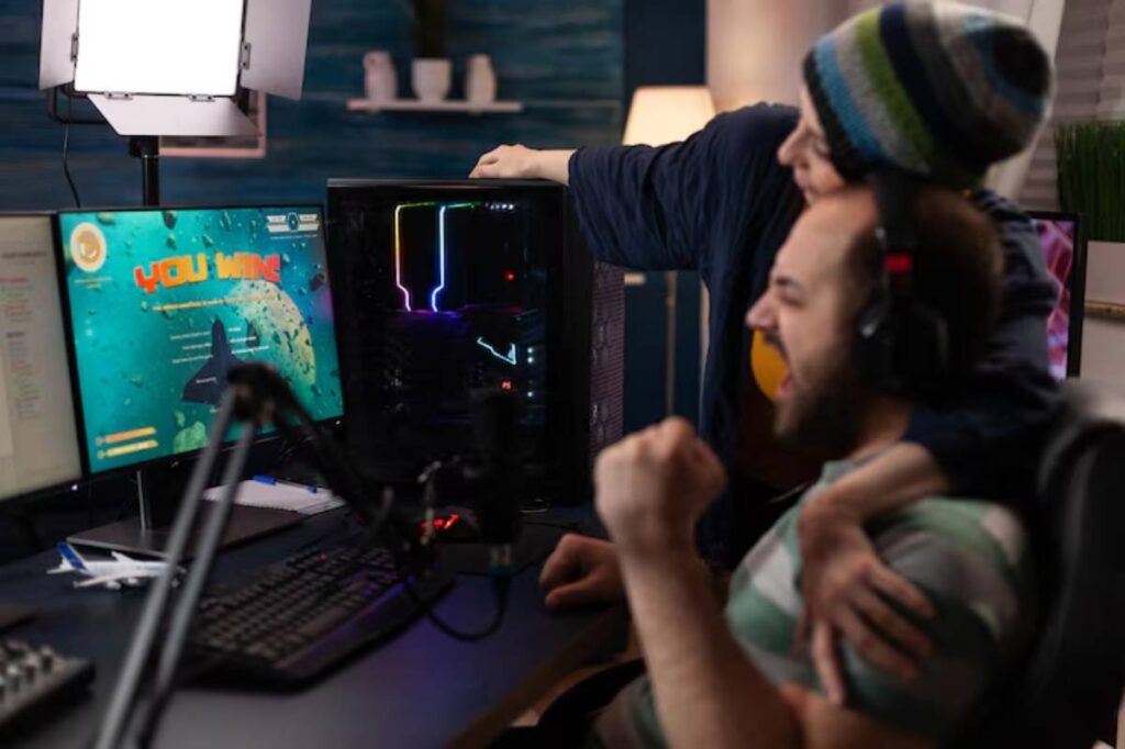 A person celebrates a gaming victory at a desk with a gaming monitor and colorful computer case, while a women hugs them from behind.