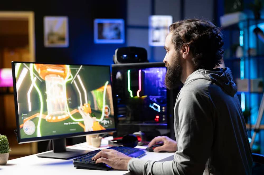 A gamer sits at a desk, focused on a vibrant video game displayed on a monitor, with a second computer screen glowing in the background.