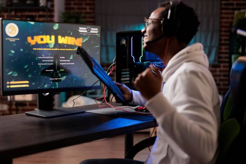 A gamer in a white hoodie celebrates a victory displayed on the screen, surrounded by gaming equipment and a stylish, modern setup.