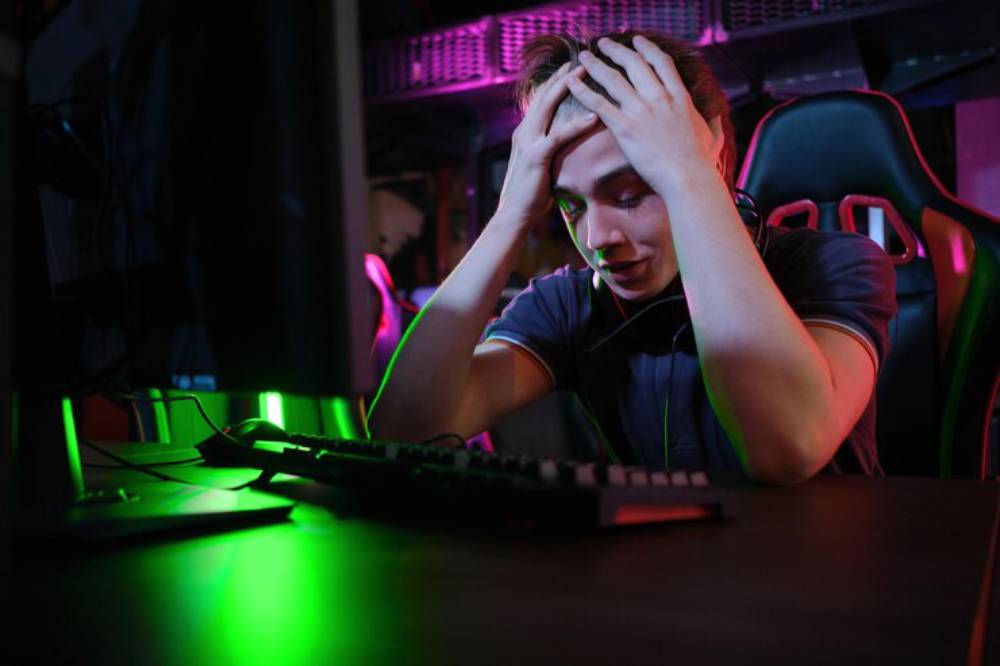 A person sits at a gaming setup with hands on their head, expressing frustration, surrounded by colorful LED lights.