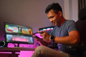 A person seated at a gaming setup, holding a console controller, with colorful monitors and ambient lighting in the background.