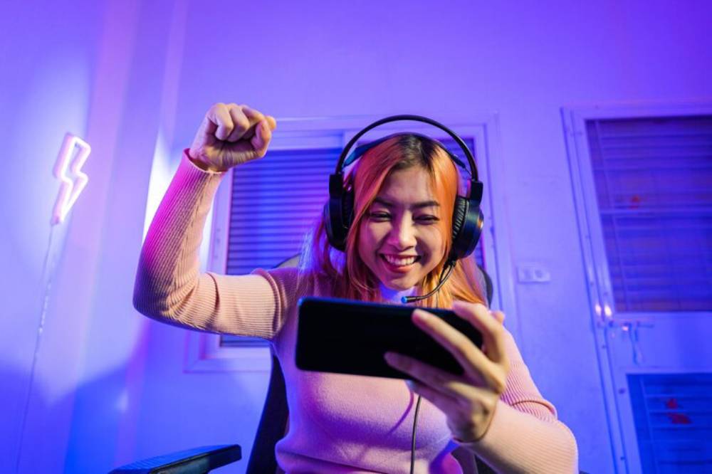 A person wearing headphones and a pink sweater is enthusiastically holding a smartphone, with a fist raised in excitement.
