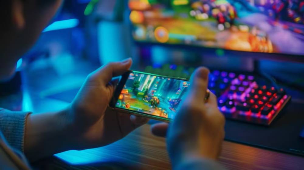 A gamer holds a smartphone playing a vibrant game, with a colorful RGB keyboard and monitor glowing in the background.