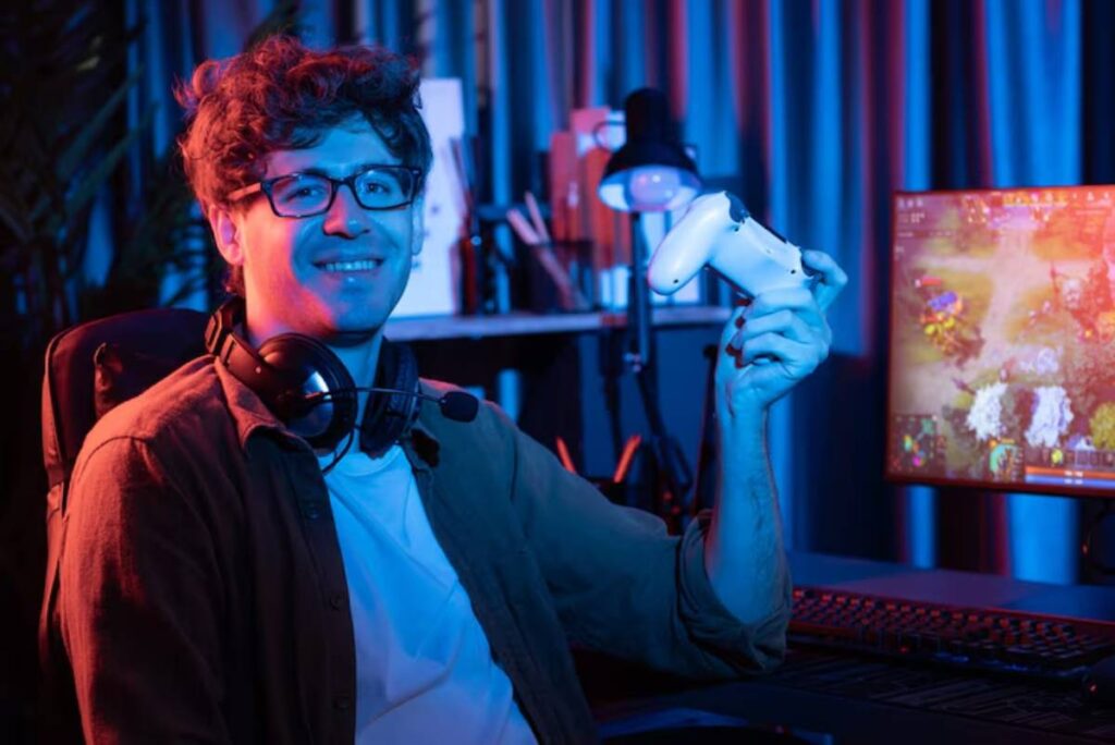 A gamer holding a game controller, wearing headphones, in a dimly lit gaming setup with a colorful screen in the background.