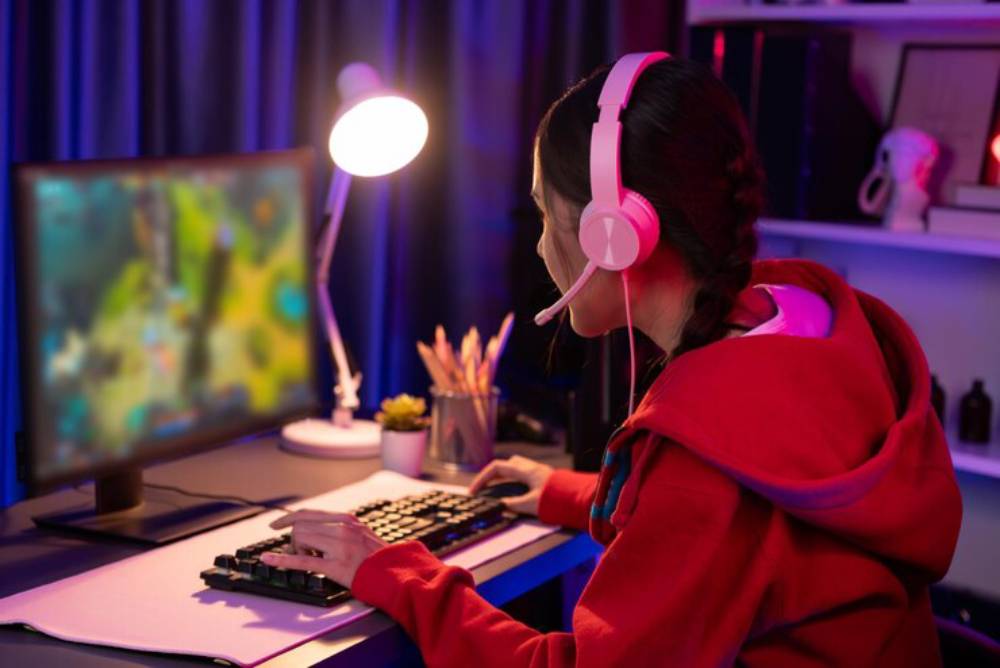 A gamer in a red hoodie uses a keyboard and mouse, focused on a colorful video game displayed on the monitor in a dimly lit room.