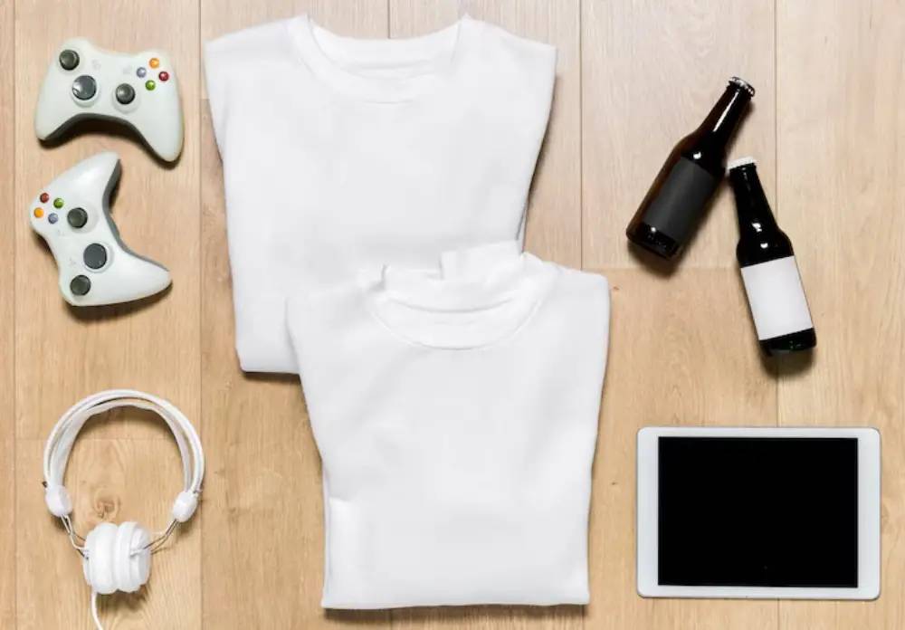 Flat lay of two white sweatshirts, gaming controllers, headphones, a tablet, and two beer bottles on a wooden surface.