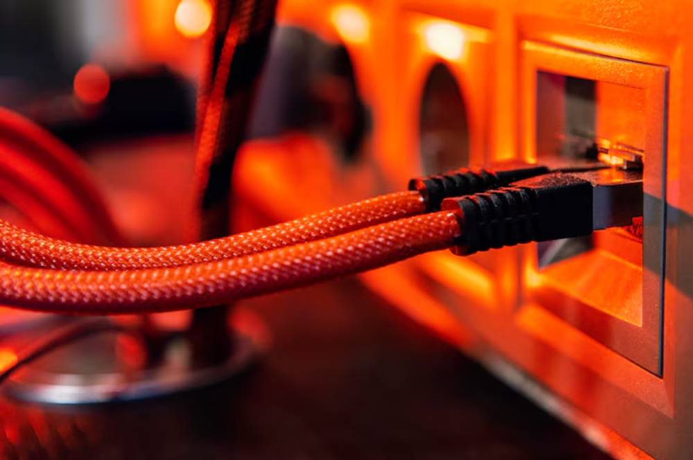 Close-up of two red braided cables connecting to an orange-lit power outlet. Background features blurred tech equipment.