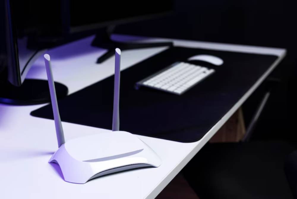 A modern white wireless router with two antennas sits on a sleek desk, beside a keyboard and mouse, against a dark background.