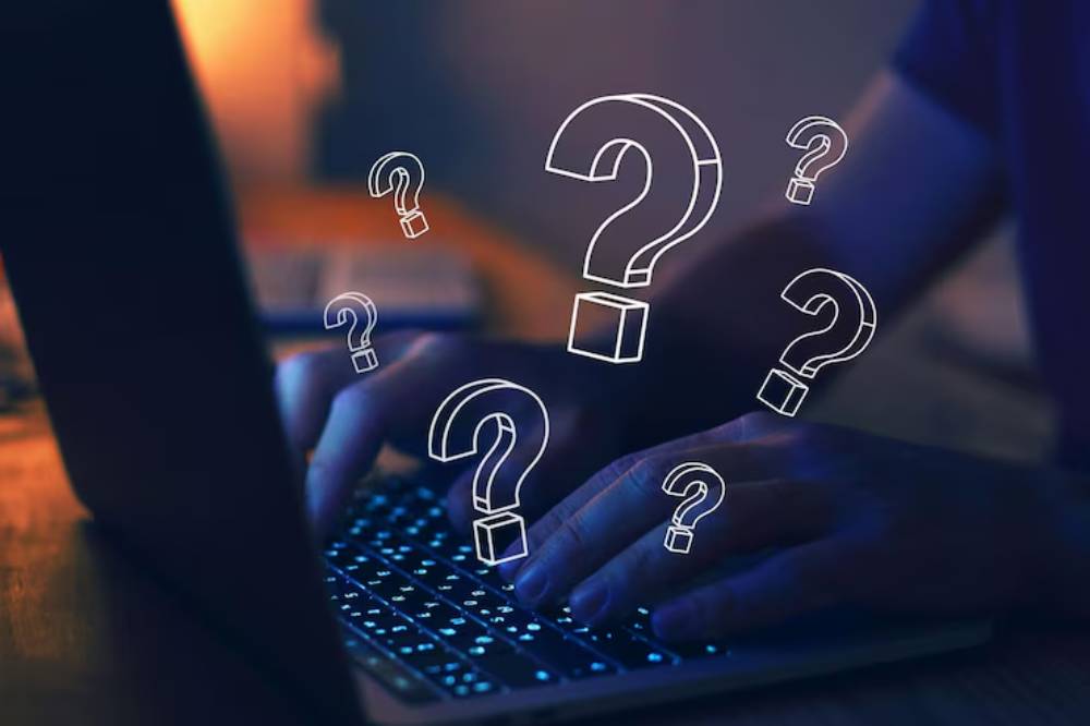 A person types on a laptop in a dimly lit room, with glowing question marks floating above, suggesting curiosity or confusion.