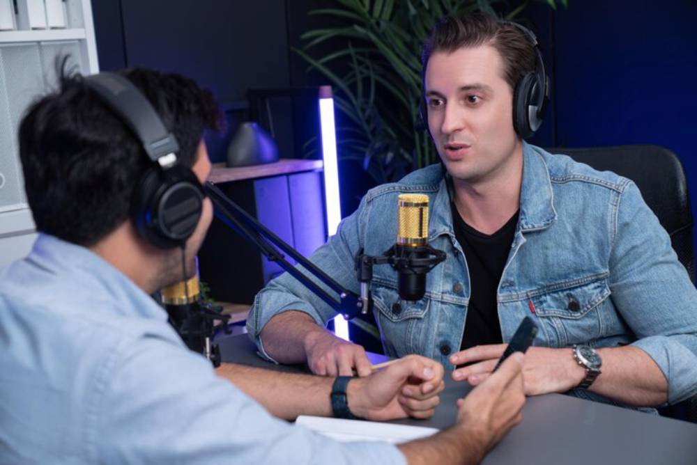 Person one is taking interview of the other  wearing headphones sit at a desk with microphones, engaged in a discussion, surrounded by modern decor.