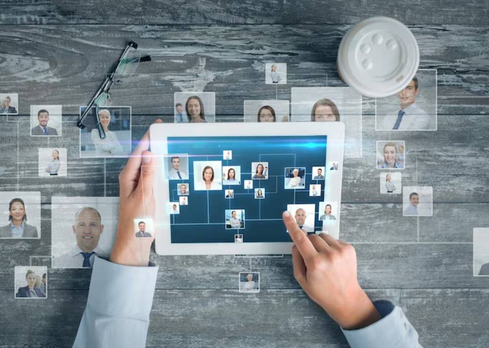 A person interacts with a tablet displaying a digital network of professional profiles, amidst a workspace with glasses and a coffee cup.