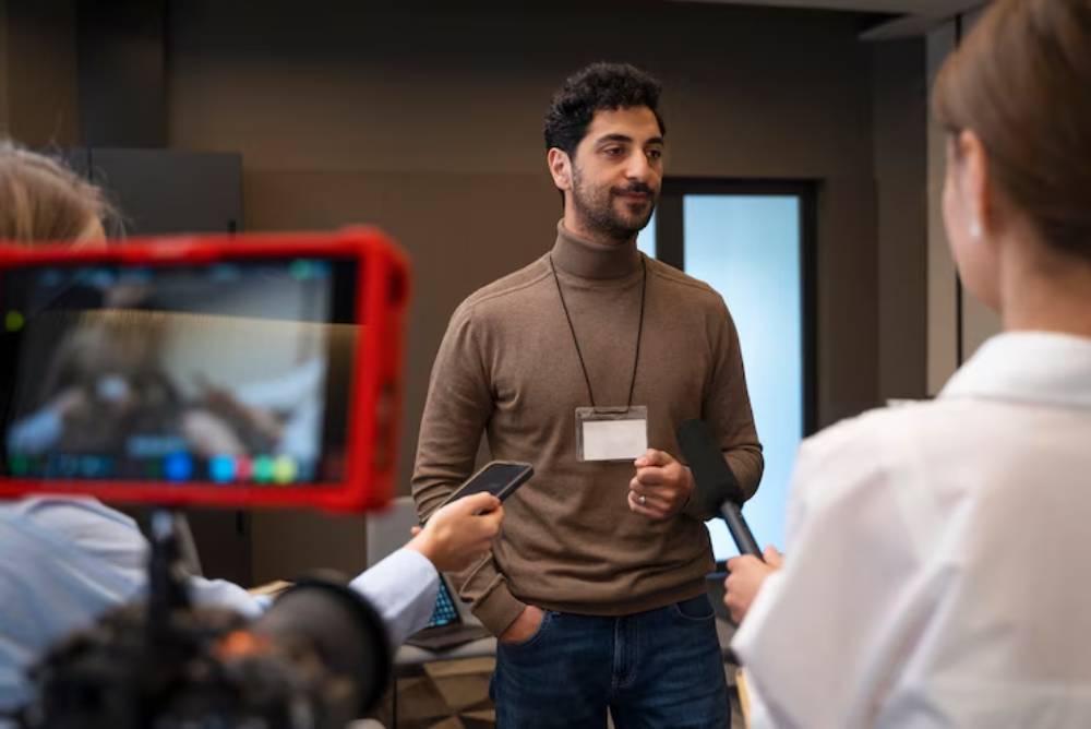 A man in a turtleneck shirt speaks during an interview, surrounded by reporters holding microphones and recording equipment.