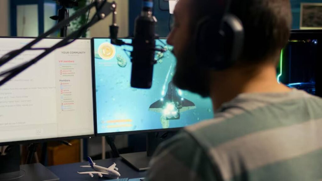 A person wears headphones and sits in front of dual monitors, one displaying a vibrant underwater game scene, with a model airplane on the desk.