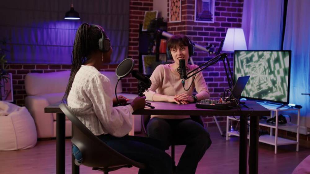 Two women sit at a table, microphones ready, in a cozy, modern studio with a warm ambiance and colorful lighting.