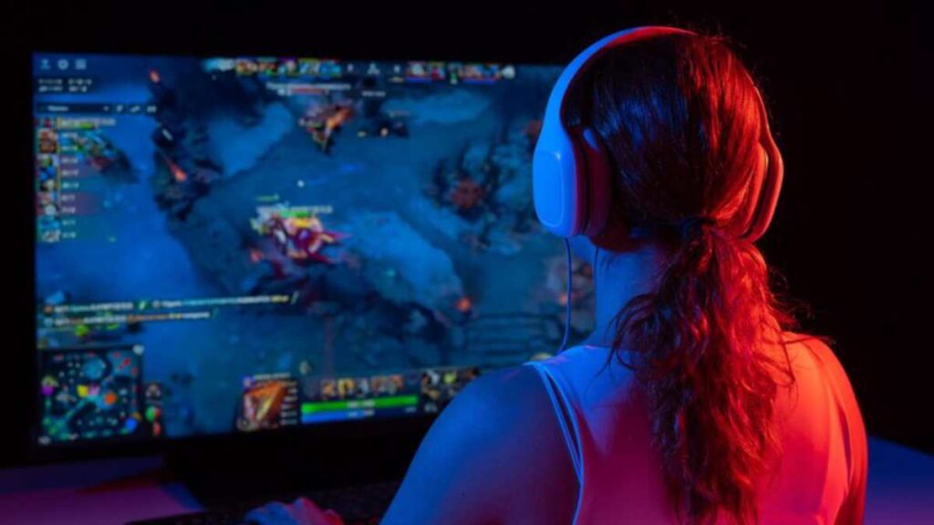 A gamer wearing headphones focuses on a colorful esports game on a large screen, illuminated in blue and red lighting.