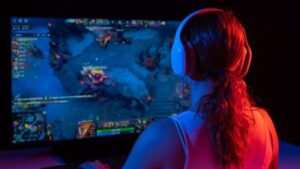 A gamer wearing headphones focuses on a colorful esports game on a large screen, illuminated in blue and red lighting.