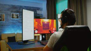 A gamer in a headset sits at a desk with dual monitors, one displaying a game and the other showing chat or streaming content.
