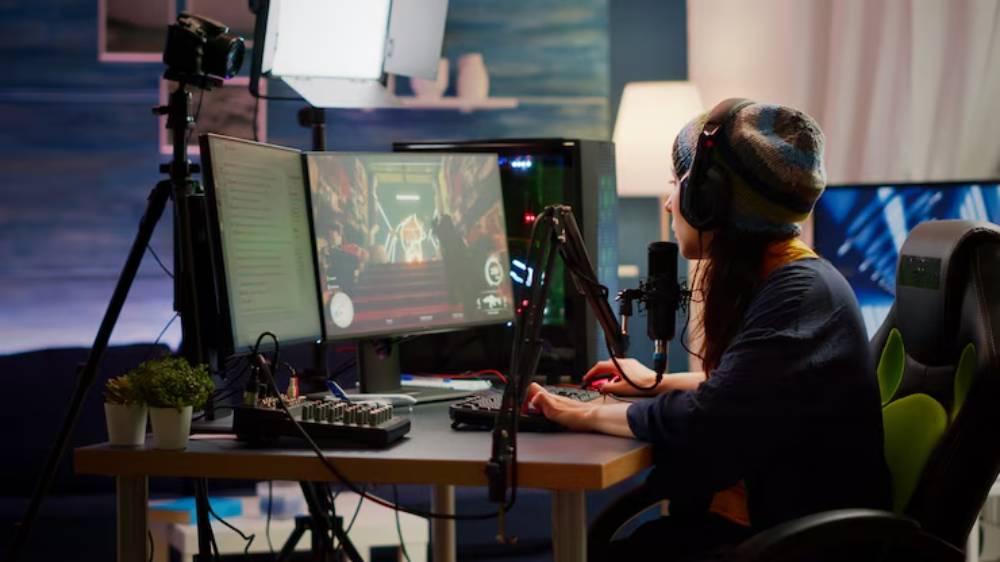 A gamer wearing headphones plays on dual monitors in a cozy setup, with a camera and lighting equipment for streaming.