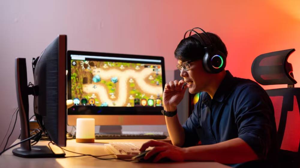 A person in a dark room wearing headphones plays a colorful video game on a computer with ambient lighting. A candle glows nearby.