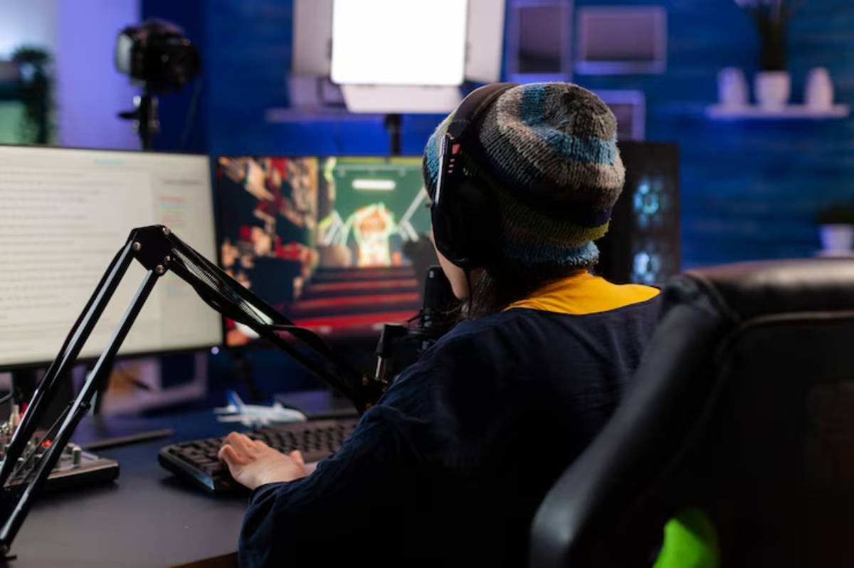 A gamer in a colorful beanie uses a keyboard and dual monitors, focused on gameplay, with studio lighting in a cozy setup.