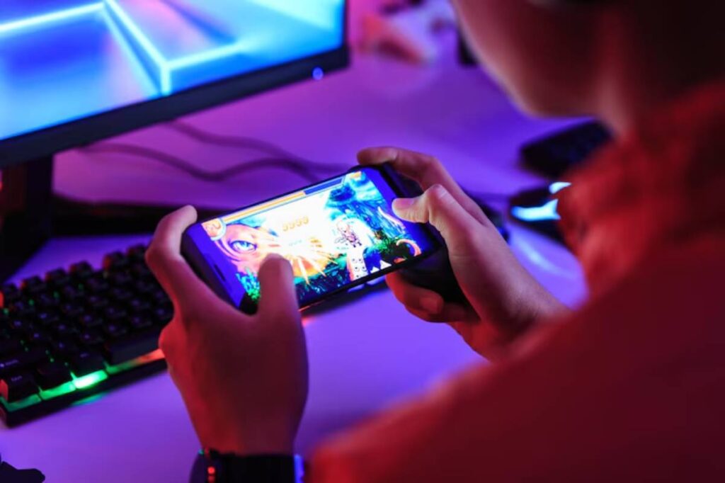 A person holds a smartphone playing a colorful game, with a backlit keyboard and monitor in a dimly lit gaming setup.