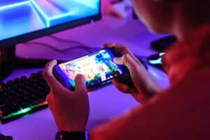 A person holds a smartphone playing a colorful game, with a backlit keyboard and monitor in a dimly lit gaming setup.