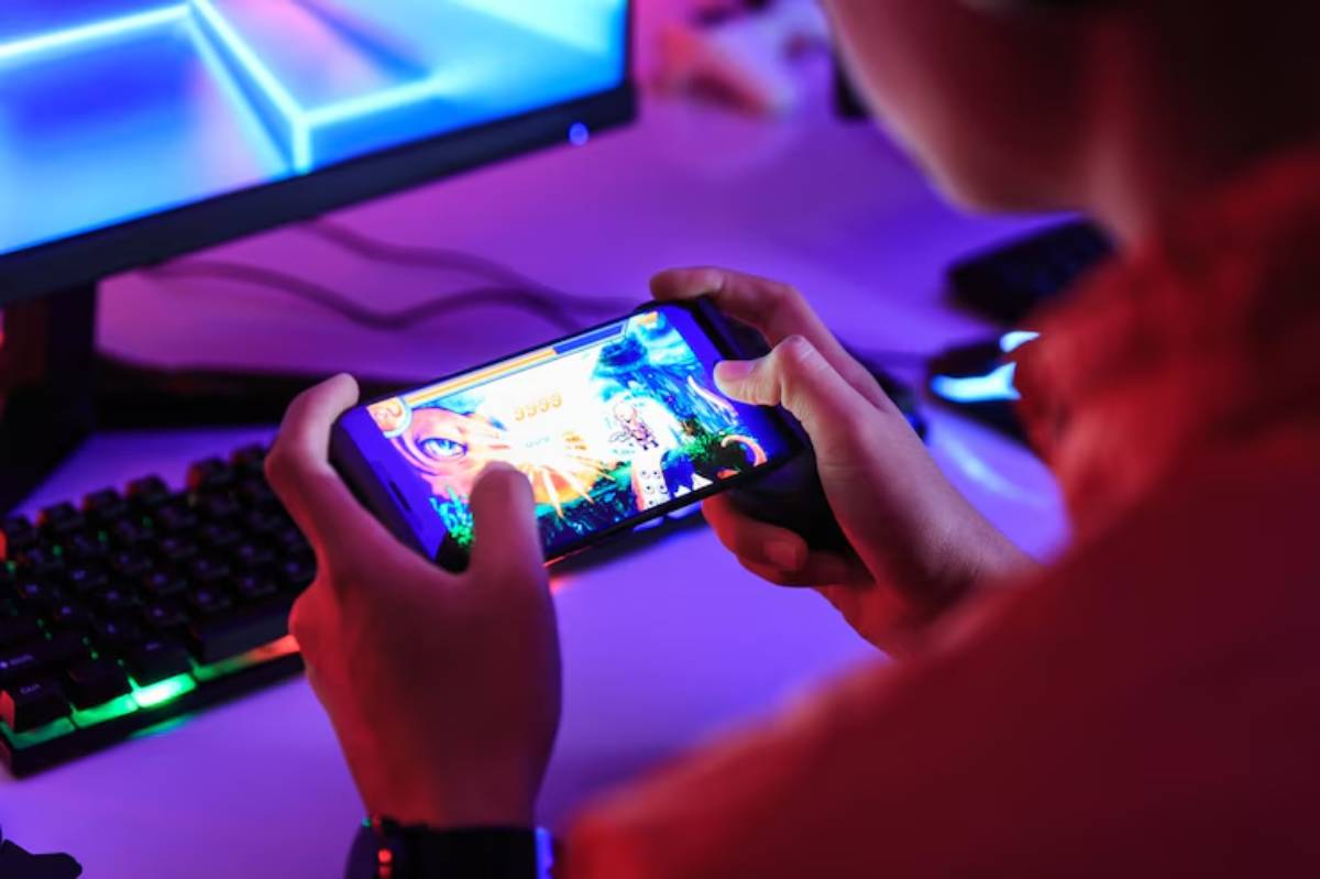 A person holds a smartphone playing a colorful game, with a backlit keyboard and monitor in a dimly lit gaming setup.