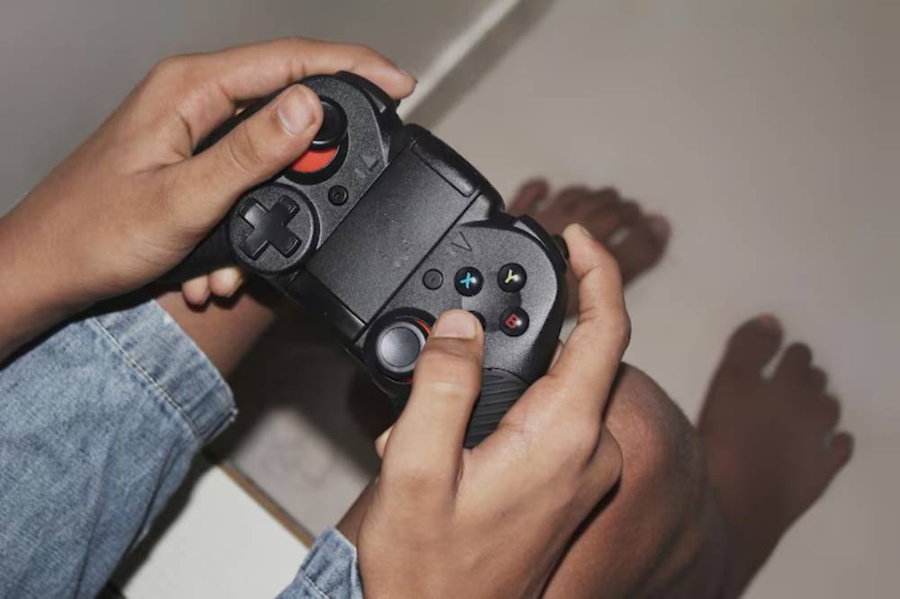 A person holds a black video game controller, with bare feet visible on the floor, focused on gameplay.