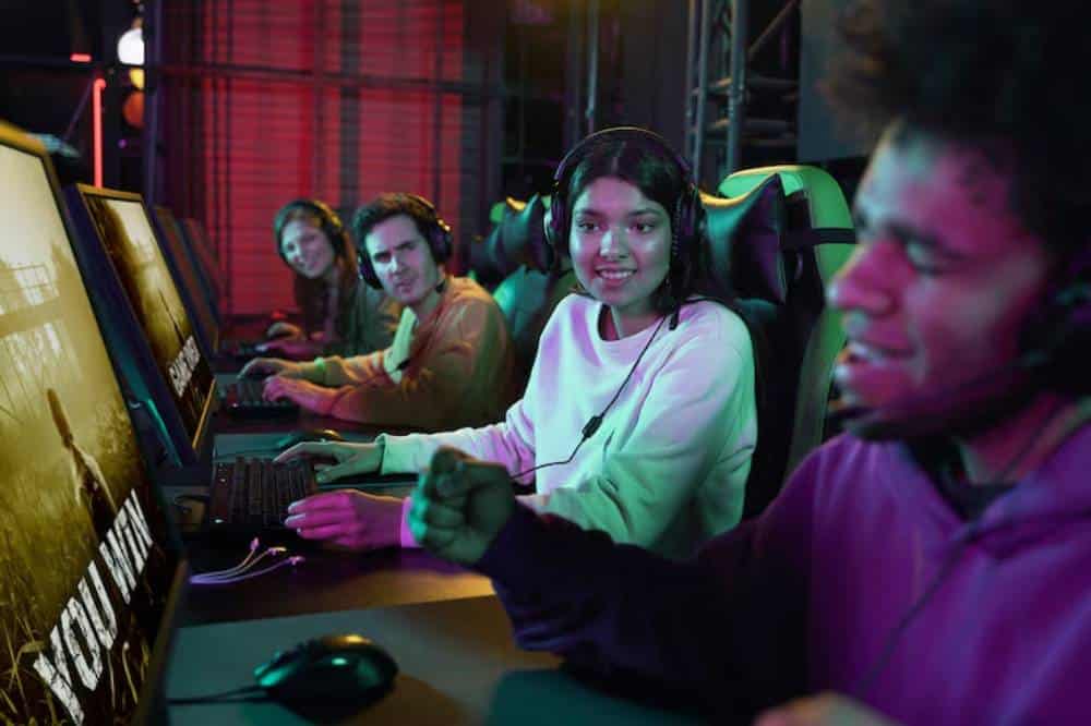 A group of gamers wearing headsets sits at computer stations, focused on their screens in a colorful, neon-lit gaming environment.