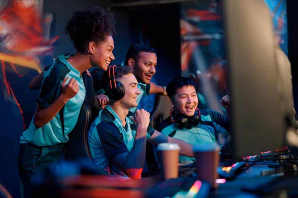 A group of excited gamers, wearing matching teal jerseys, cheer enthusiastically around a computer setup in a vibrant gaming environment.