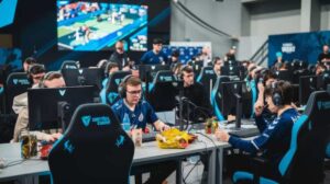 A crowded esports event featuring gamers seated at desks with monitors, headsets, and snacks, focused on a large screen displaying gameplay.