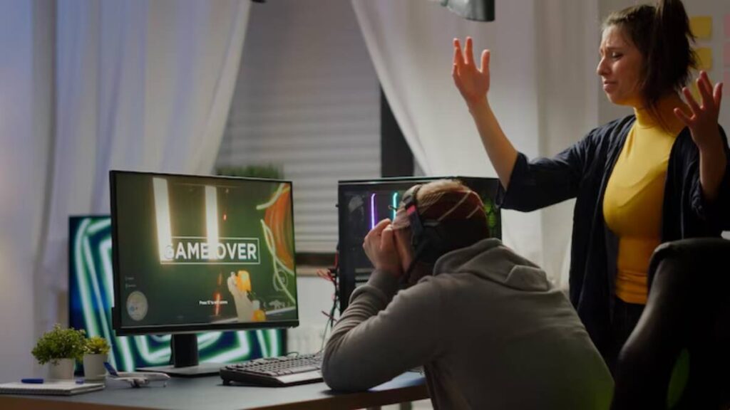 A gamer sits at a computer, facepalming in frustration, while a person in a yellow shirt gestures animatedly behind them.