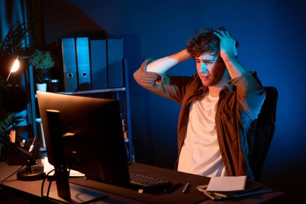 A person sits at a desk in a dimly lit room, holding their head in stress, surrounded by a computer and office supplies.