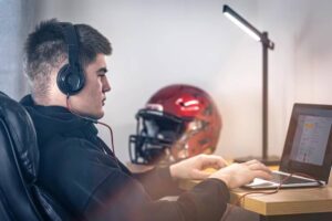 A person wearing headphones sits at a desk using a laptop, with a red football helmet prominently displayed nearby.